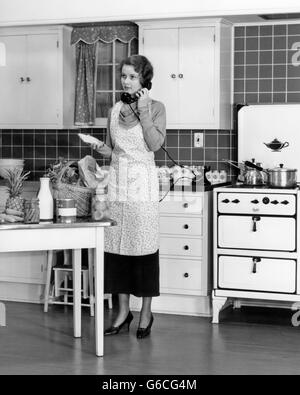 1930 FEMME DANS LA CUISINE L'ÉPICERIE SUR LA TABLE TALKING ON TELEPHONE Banque D'Images