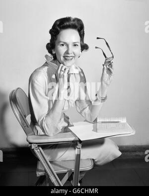 1950 SMILING WOMAN SITTING IN CLASSROOM PRÉSIDENT DE HOLDING GLASSES RESTING CHIN ON HAND LOOKING AT CAMERA Banque D'Images