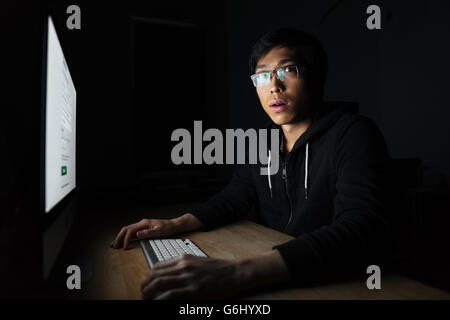 Jeune homme asiatique surpris dans les verres travailler avec ordinateur de bureau sombre Banque D'Images