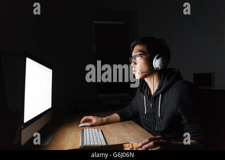 Bel asiatique jeune homme à lunettes et casque à l'aide d'ordinateur et de manger la pizza au bureau sombre Banque D'Images