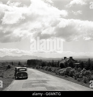 Dans les années 1950, historique, une dame se tient près de sa voiture, une Austin de construction britannique, garée sur le côté d'une route de campagne déserte, l'Irlande. Banque D'Images