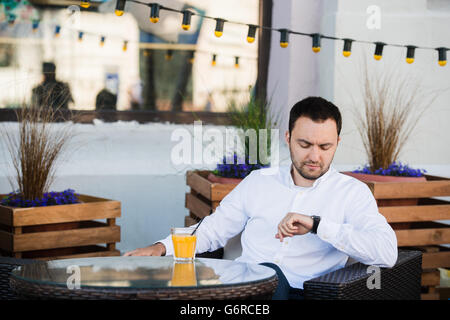 Handsome businessman est en attente de client en cafétéria. Il est touchant et montre à la recherche avec l'anticipation. L'homme est assis à l'extérieur de la table et sourit. Il boit du jus d'orange Banque D'Images