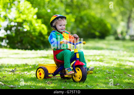 Cute boy wearing safety helmet équitation son tricycle en parc d'été ensoleillé. Les enfants circuler à bicyclette. Premier vélo pour petit enfant. Banque D'Images