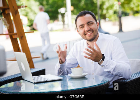 Young handsome man smiling et boire du café dans un restaurant en bord de mer. Il est heureux, looking at camera Banque D'Images