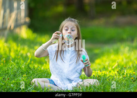 Little girl blowing bubbles assis sur l'herbe. Banque D'Images