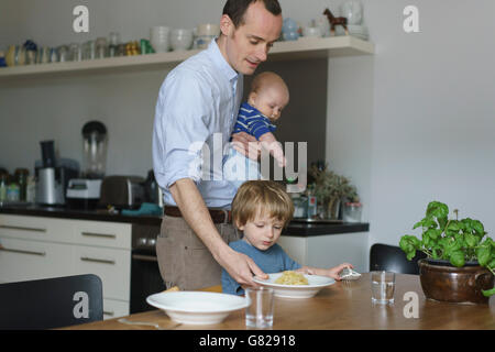 Père exerçant son tout-petit tout en donnant de la nourriture à son fils à table à manger Banque D'Images