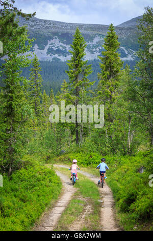 Deux jeunes enfants faire du vélo sur route forestière, parmi les grands arbres et montagnes. Petites Personnes dans le cadre. Banque D'Images