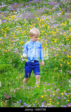 Garçon marche dans une prairie remplie de fleurs de différentes couleurs, tenant un bouquet dans sa main. Porter des vêtements formels, cravate. Banque D'Images