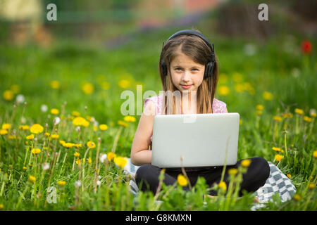 Petite fille dans les écouteurs avec ordinateur portable assis sur l'herbe dans le parc. Banque D'Images