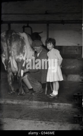 Années 1930, photo historique montrant une jeune fille debout dans un enclos à côté d'un agriculteur qui est traire une vache à la main, en Angleterre. Banque D'Images