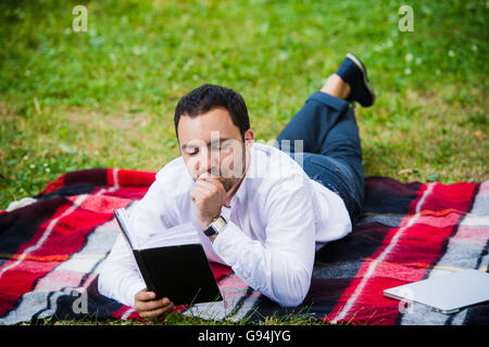Beau jeune businessman reading book lying in the park Banque D'Images