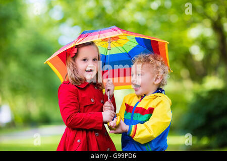 Petite fille jouant dans le parc de l'été pluvieux. Parapluie arc-en-ciel avec l'enfant, manteau imperméable et des bottes de saut en flaque sous la pluie. Banque D'Images