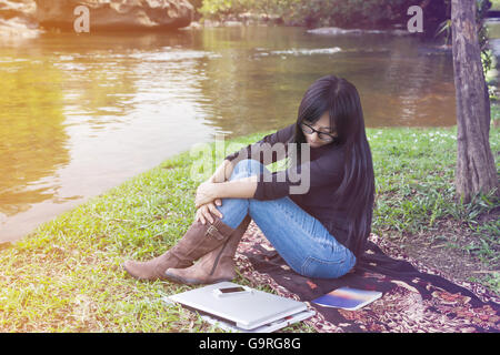 Girl sitting on grass field at Riverside avec ordinateur portable et smartphone, fille ou femme looking at smartphone ou appel téléphonique en attente je Banque D'Images