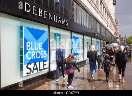 Des problèmes de mise au point floue, les personnes consommateurs chiffres passant de l'été de la Croix Bleue à signes de vente magasin Debenhams dans Piccadilly, Manchester, Royaume-Uni. Vente et réductions, promotions & offres spéciales, articles de dégagement, remise, prix, promotion, commerce de détail, vie quotidienne, scènes de rue en dehors de Debenhams department store. Banque D'Images