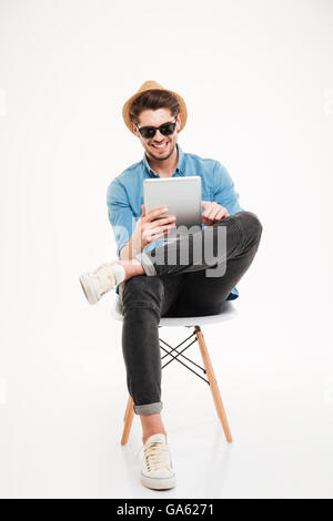 Cheerful attractive young male wearing glasses sitting and holding tablet isolé sur fond blanc Banque D'Images