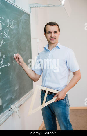 Jeune enseignant ou étudiant holding triangle qui pointe à l'ardoise avec formule, à la recherche d'appareil photo et smiling Banque D'Images