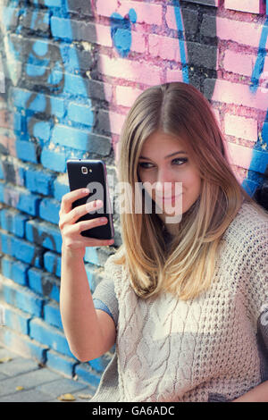 Jeune femme blonde, la navigation sur Internet avec le téléphone, et souriant tout en restant assis sur le sol contre le mur de brique avec graffiti Banque D'Images