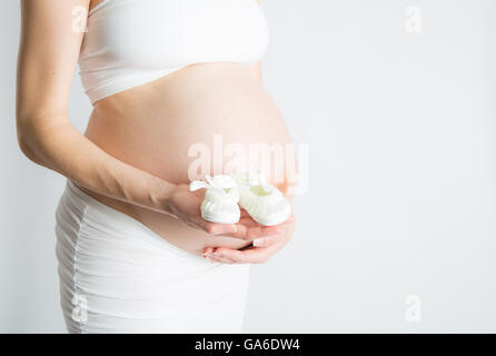 Une femme enceinte est titulaire d'une paire de chaussures de bébé en face de son ventre enceinte. Banque D'Images
