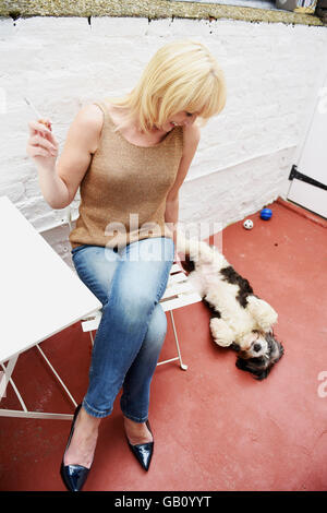 Woman enjoying a cigarette dans sa cour avec son chien de compagnie roulant sur son dos Banque D'Images