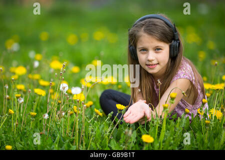 Peu cute girl in headphones assis sur l'herbe verte dans le parc. Banque D'Images