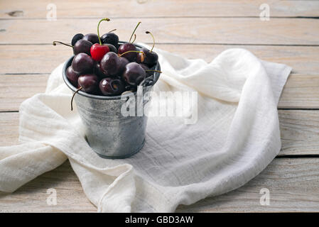 Les cerises mûres dans un seau en métal sur une nappe et une table en bois Banque D'Images