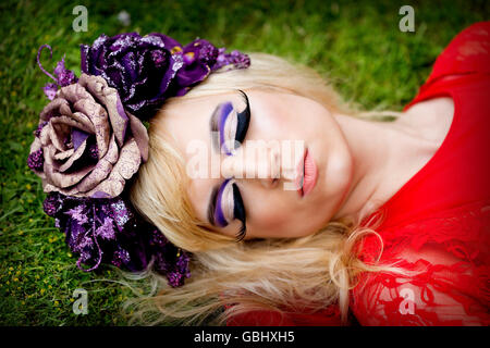 Blonde modèle, traits délicats, la couronne de fleurs de mauve, ailé, d'eyeliner cils énorme, robe en dentelle rouge, couché sur l'herbe de couchage Banque D'Images