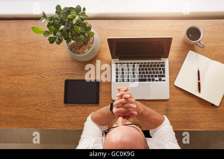 Vue d'en haut photo de l'homme assis à son bureau de travail et de pensée. Ordinateur portable, tablette numérique, journal, tasse à café et plante en pot sur le sol en bois Banque D'Images