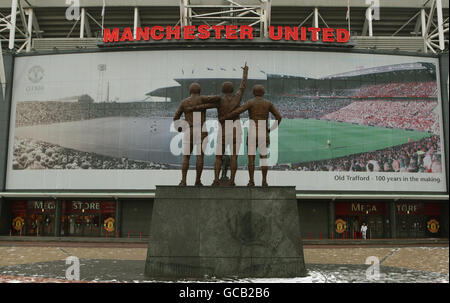 La statue de la Trinité-Uni de Best, Law et Charlton fait face à une affiche sur le stand est du stade de Manchester United montrant un montage et une version ancienne et nouvelle du terrain à Old Trafford, Manchester. Banque D'Images