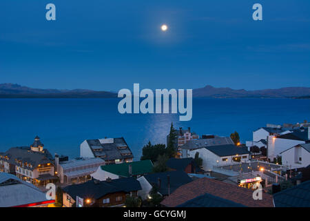 La Lune se levant au-dessus d'un lac au-dessus de la ville de toits montrant quelques lumières de la ville jaune sur le fond bleu, Bariloche, Argentine Banque D'Images