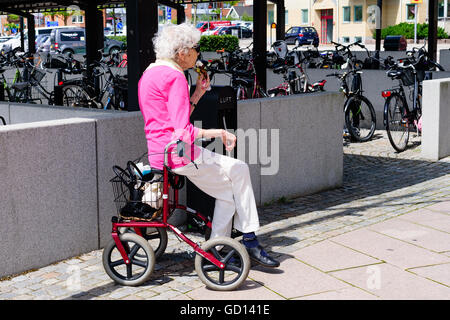 Ronneby, Suède - Juillet 9, 2016 : Senior woman sitting on walker la consommation de crème glacée. Stationnement pour vélos dans l'arrière-plan. Banque D'Images