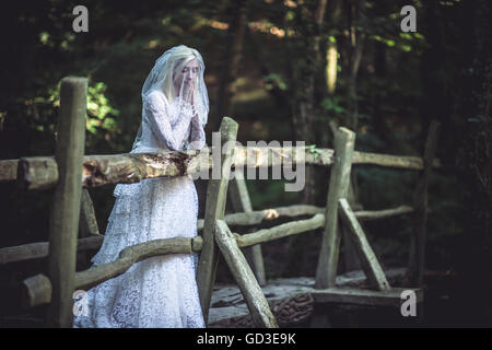 Une jeune femme blonde girl wearing une ancienne robe de mariée blanche robe debout sur un pont en bois rustique dans un bois un soir d'été,, UK Banque D'Images