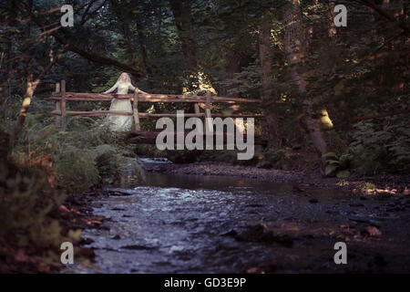 Une jeune femme blonde girl wearing une ancienne robe de mariée blanche robe debout sur un pont en bois rustique dans un bois un soir d'été,, UK Banque D'Images