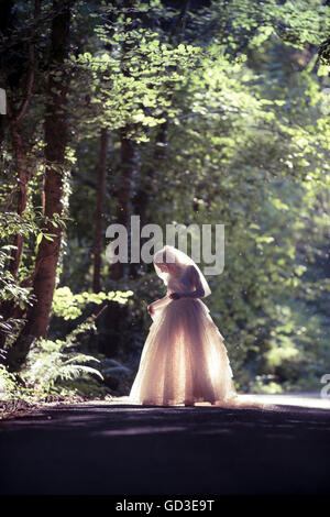 Une jeune femme blonde girl wearing une ancienne robe de mariage blanc rétroéclairé redingote debout dans une forêt un soir d'été,, UK Banque D'Images