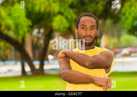 Homme portant chemise jaune bras étirement dans le parc entouré d'herbe verte et des arbres, le concept de formation Banque D'Images