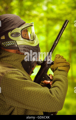 Closeup portrait man wearing jacket, vert et noir masque facial de protection de l'article profil en angle avec arme, fond de forêt, concept paintball Banque D'Images