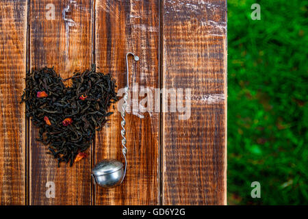 Crépine vintage près de feuilles sèches de thé noir faites en coeur sur table en bois dans le jardin et sur la nature de fond. concept de thé le thé. Banque D'Images