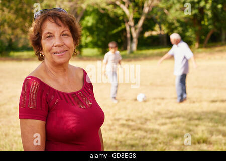 Portrait de confiant et heureux grand-mère, smiling at camera, tandis que son petit-fils et mari jouer au football en arrière-plan. Banque D'Images