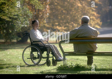 Femme assise dans un fauteuil roulant, homme assis sur un banc de parc Banque D'Images