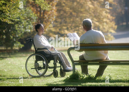 Femme assise dans un fauteuil roulant, homme assis sur un banc de parc avec un livre Banque D'Images