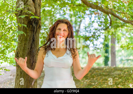 Close up of young woman smiling classy busty à l'appareil photo tout en faisant bienvenue signe shaka contre green garden background with copy space Banque D'Images