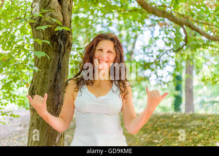 Close up of young woman smiling classy busty à l'appareil photo tout en faisant bienvenue signe shaka contre green garden background with copy space Banque D'Images