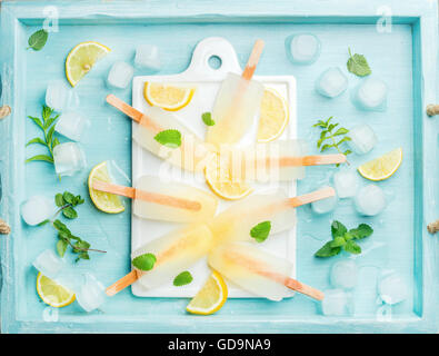 Sucettes glacées au citron sur conseil en céramique blanche servi avec des rondelles de citron, les cubes de glace et de feuilles de menthe sur fond bleu turquoise, top v Banque D'Images