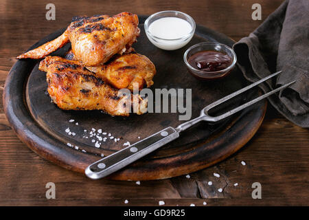 Grillades de viande de poulet ailes et les pattes servi sur planche de bois rond avec deux sauces, sel et vintage gris sur fourchette à viande Banque D'Images