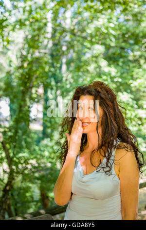 Close up of young woman smiling classy busty à l'appareil photo tout en couvrant sa bouche avec la main contre green garden background with copy space Banque D'Images