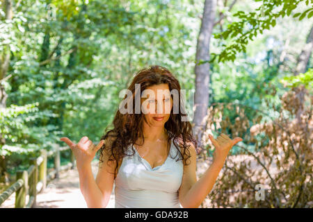 Close up of young woman chic busty tout en essayant de faire bienvenue signe shaka contre green garden background with copy space Banque D'Images