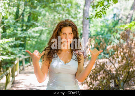 Close up of young woman smiling classy busty à l'appareil photo tout en faisant bienvenue signe shaka contre green garden background with copy space Banque D'Images