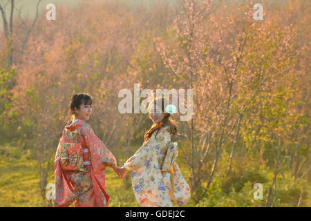 Deux smiling women holding hands in cherry blossom verger portant des vêtements traditionnels japonais Banque D'Images