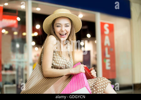 Portrait of young woman wearing hat dans centre commercial, serrant étroitement avec les achats de sacs, riant joyeusement Banque D'Images