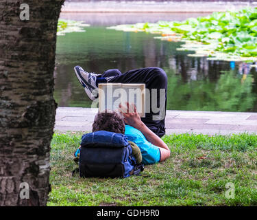 Loisirs et détente, l'homme étendu sur l'herbe et lecture réserve en parc public à côté d'un étang Banque D'Images