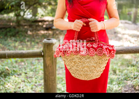 Les mains tenant un sac dans un style rustique faite de raphia et de tissu rouge avec des fleurs blanches, la femme vêtue d'une robe fourreau rouge attend nerveusement dans un parc Banque D'Images